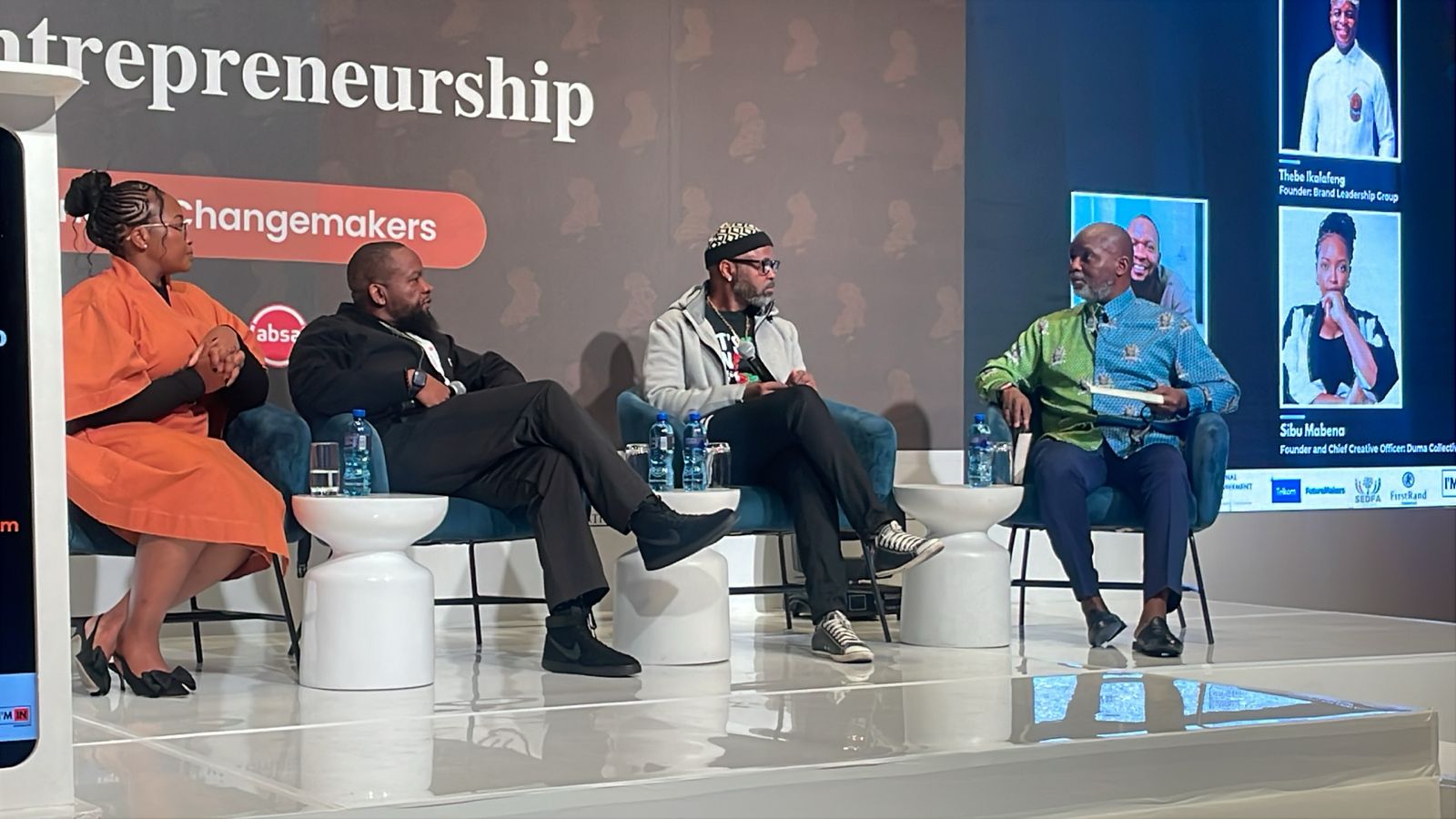 A vibrant African entrepreneurship event at a modern venue in Melrose, with panelists on a stage in a fireside chat setup. Attendees are engaged, with warm lighting and subtle branding elements of business growth and networking.