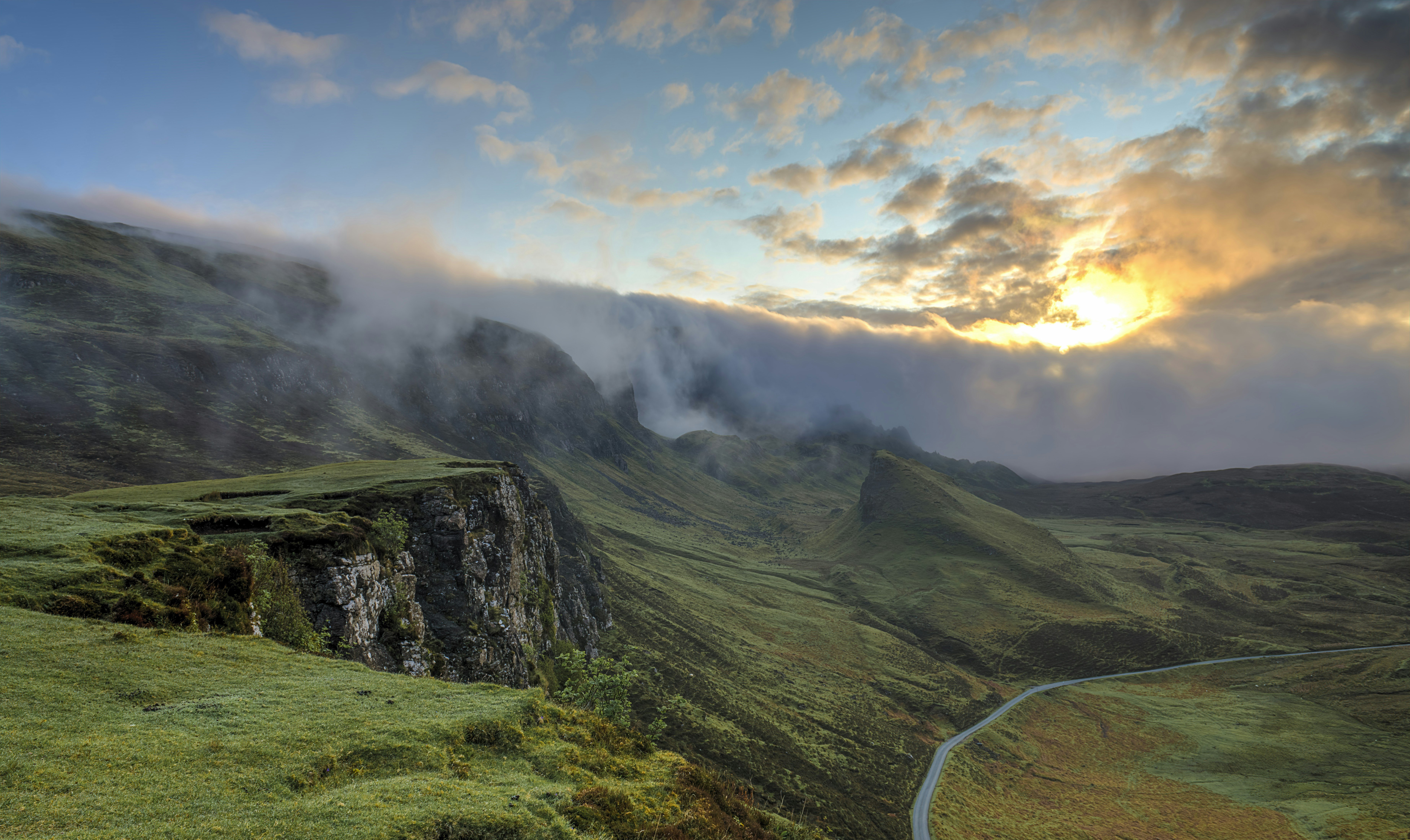 A breathtaking landscape of rolling green hills and rugged cliffs, shrouded in mist, with the sun breaking through dramatic clouds at dawn or dusk.