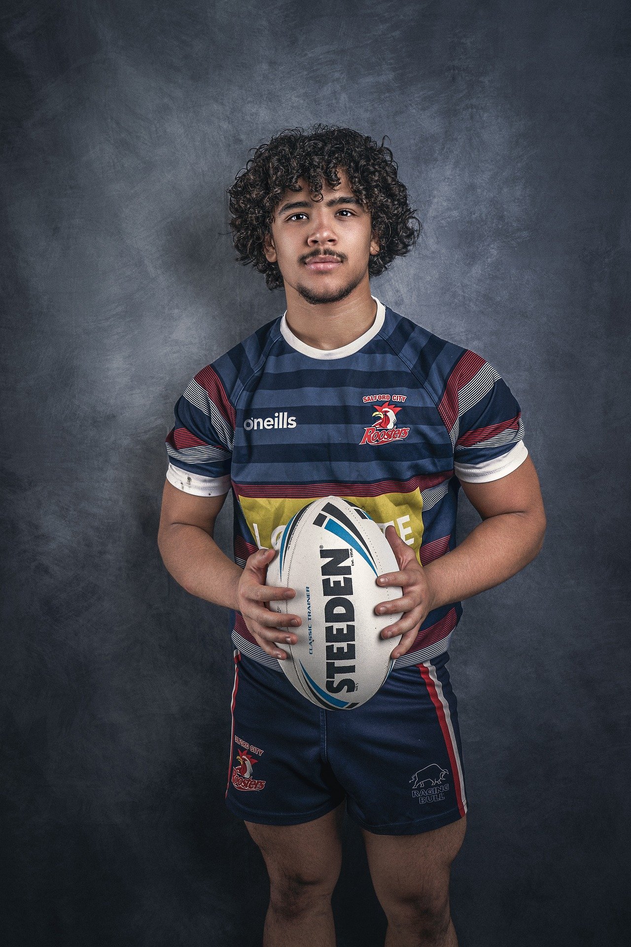 The image shows a young rugby player wearing a "Salford City Roosters" jersey, holding a Steeden rugby ball, standing in a portrait pose.